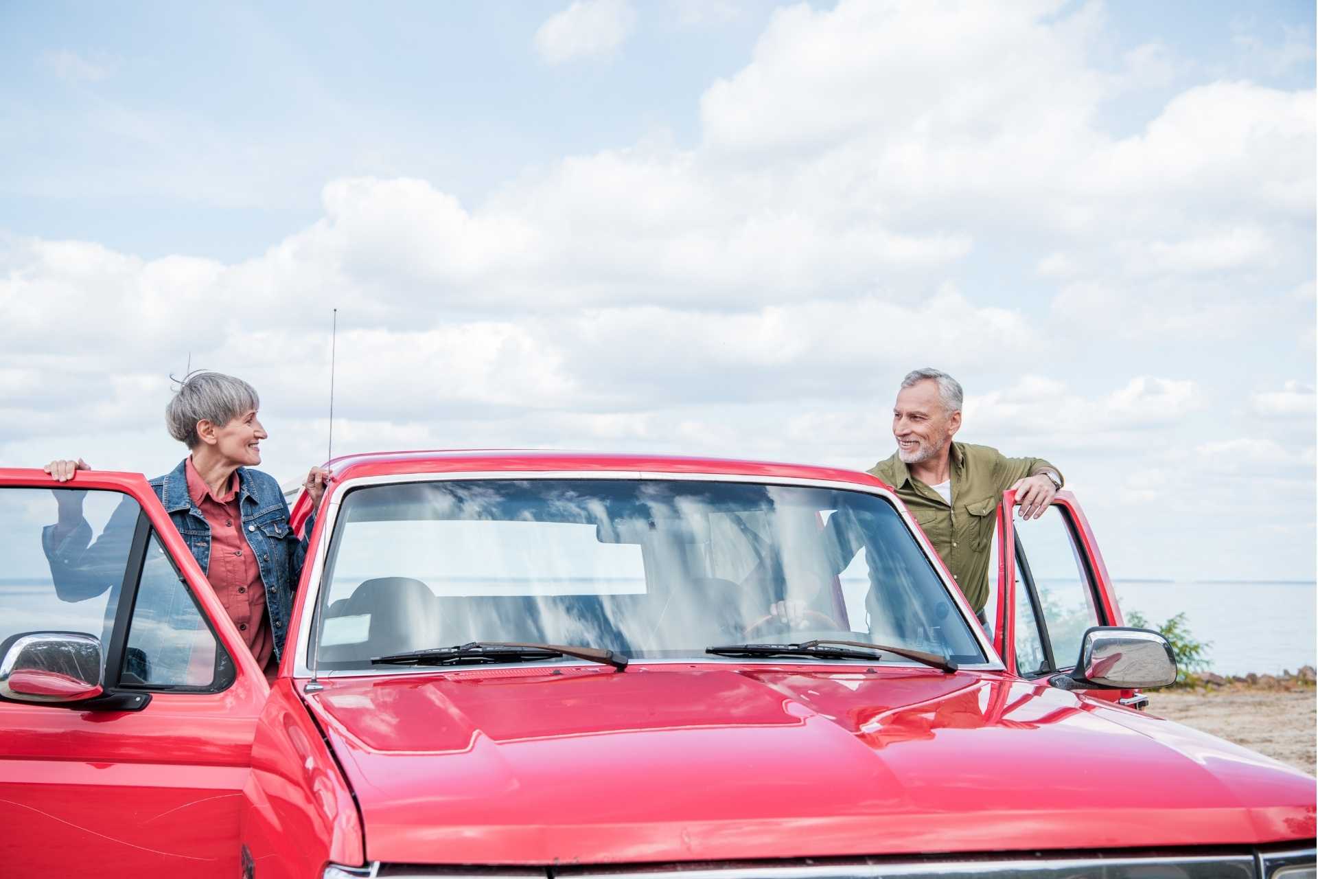 smiling senior couple of tourists standing near red car and looking at each other at beach.
